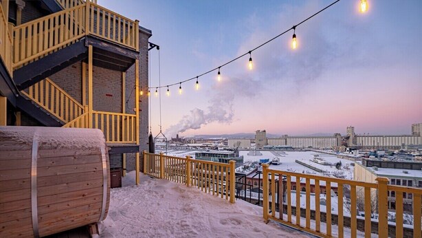 A snowy outdoor area with a wooden barrel, a yellow railing, and a view of a cityscape with buildings and a bridge in the background. - Remparts 2 Bed w Private terrace & sauna (Québec)