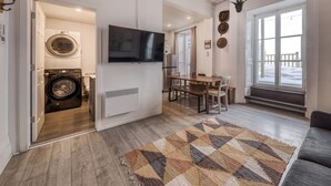 A modern living room with a flat-screen TV, a dining area with a wooden table and chairs, a large window with a view of the outdoors, and a laundry area with a washing machine and dryer. - Remparts 2 Bed w Private terrace & sauna (Québec)