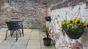Une terrasse avec une table et des chaises, un pot de fleurs et des jardinières suspendues, le tout adossé à un mur de briques.