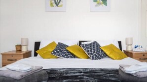Un lit bien fait, avec une literie blanche et deux coussins décoratifs, l’un jaune et l’autre orné d’un motif géométrique noir. Au-dessus du lit, sur le mur, on peut voir deux reproductions encadrées de motifs botaniques.