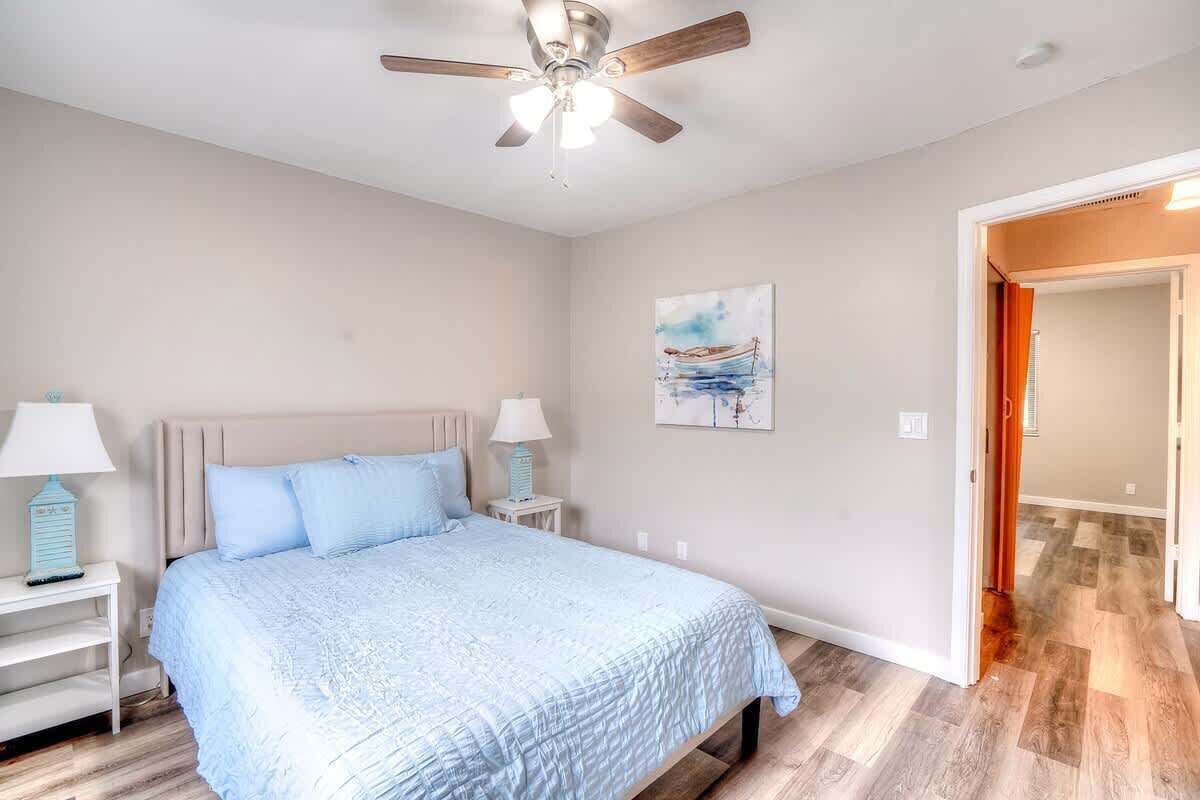 A bedroom with a bed, nightstands, a ceiling fan, a painting, and a wooden floor.