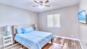 A bedroom with a bed, a ceiling fan, a window with blinds, and a wall-mounted TV.