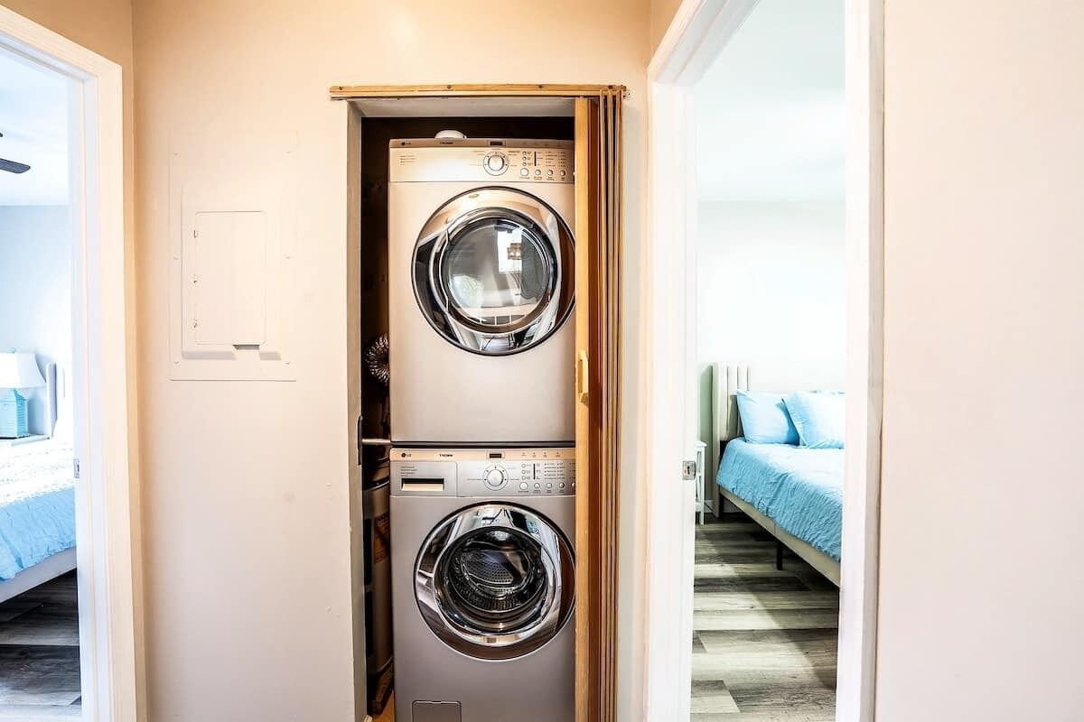 Un cuarto de lavandería con dos lavadoras de carga frontal, una cama con ropa de cama azul y piso de madera.