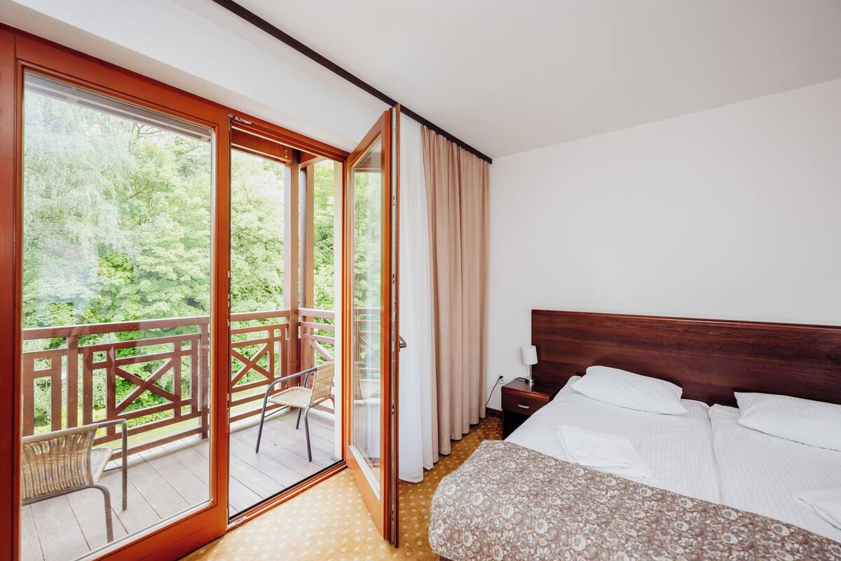 A hotel room with a bed, a wooden headboard, a nightstand, a chair, and a balcony with a view of greenery.