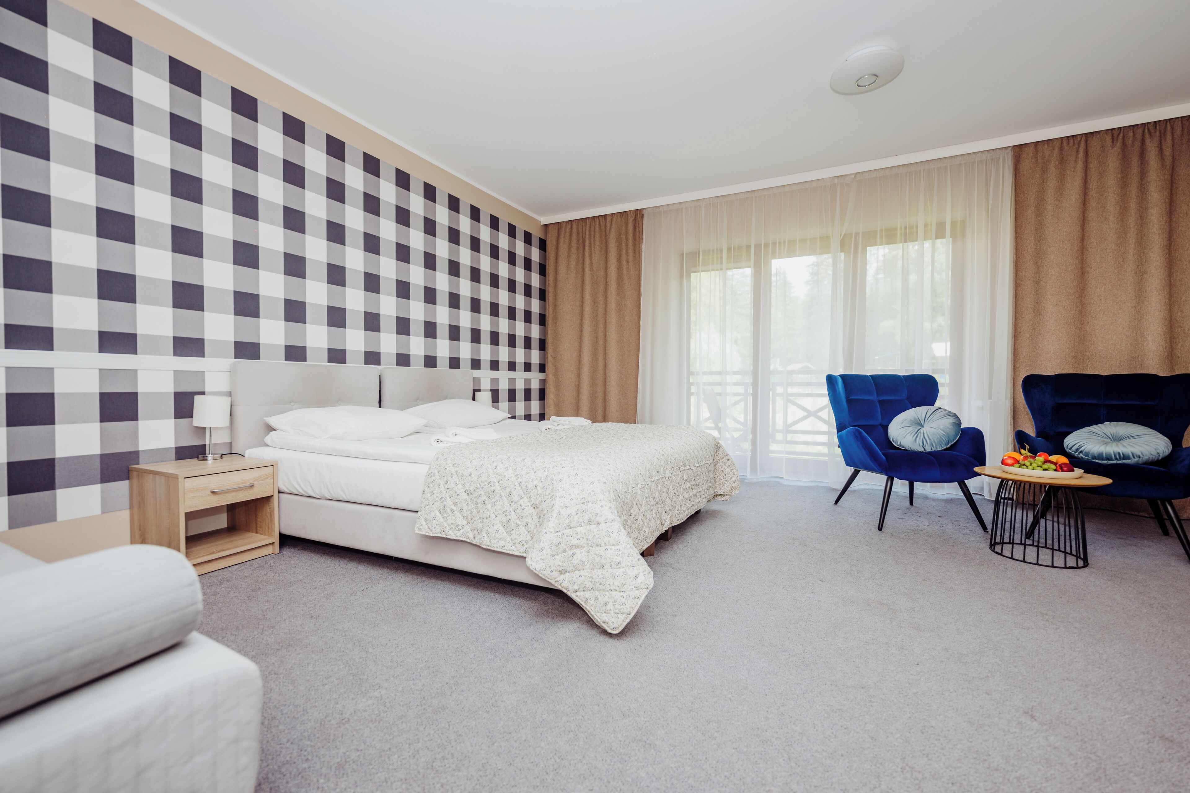 Une chambre d’hôtel avec un mur à carreaux, un lit recouvert d’une couverture blanche, un fauteuil bleu, une petite table avec des fruits et une fenêtre avec des rideaux.