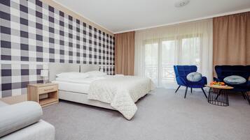 Une chambre d’hôtel avec un mur à carreaux, un lit recouvert d’une couverture blanche, un fauteuil bleu, une petite table avec des fruits et une fenêtre avec des rideaux.