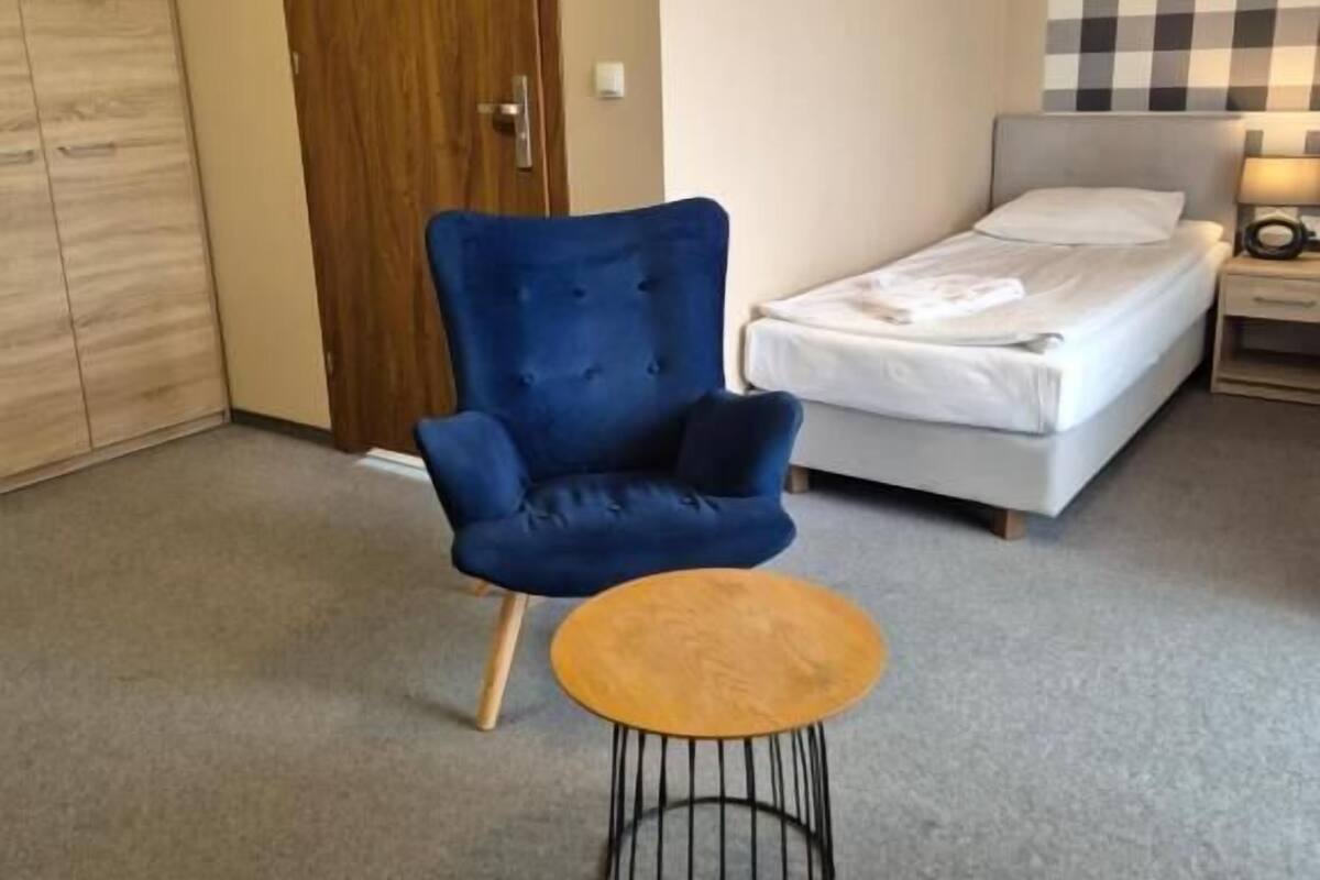 A room with a bed, a blue armchair, a small round table, a wooden wardrobe, and a checkered wall.