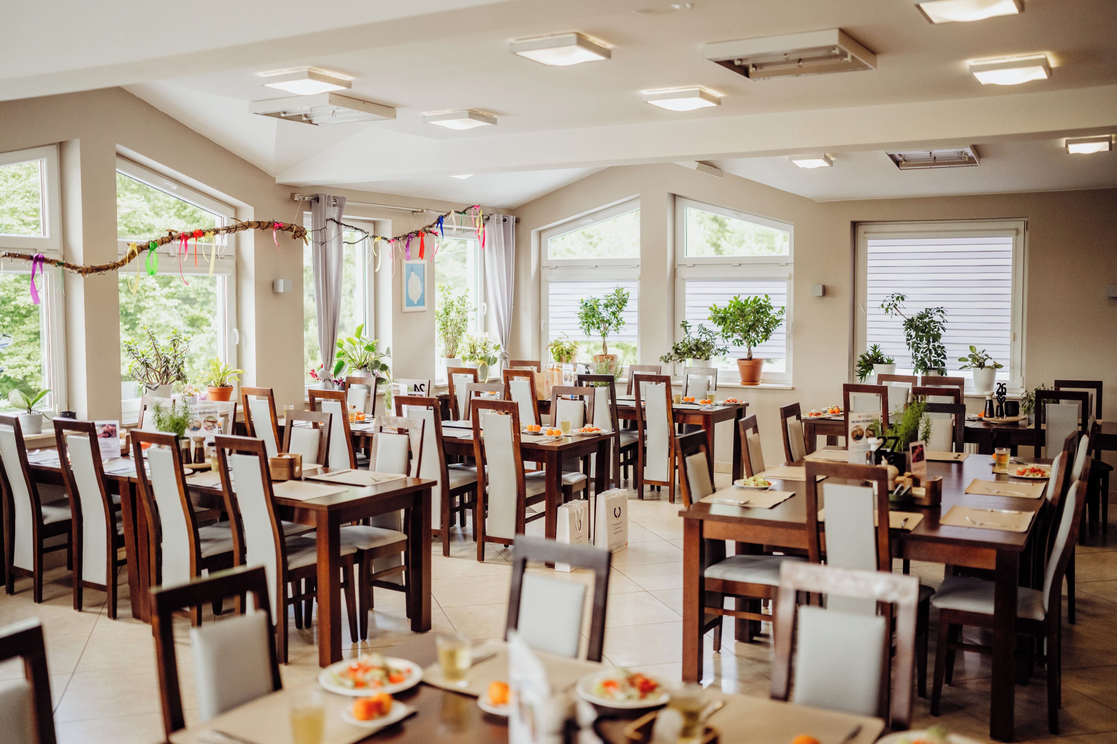 A spacious dining area with multiple tables and chairs, large windows, and potted plants.