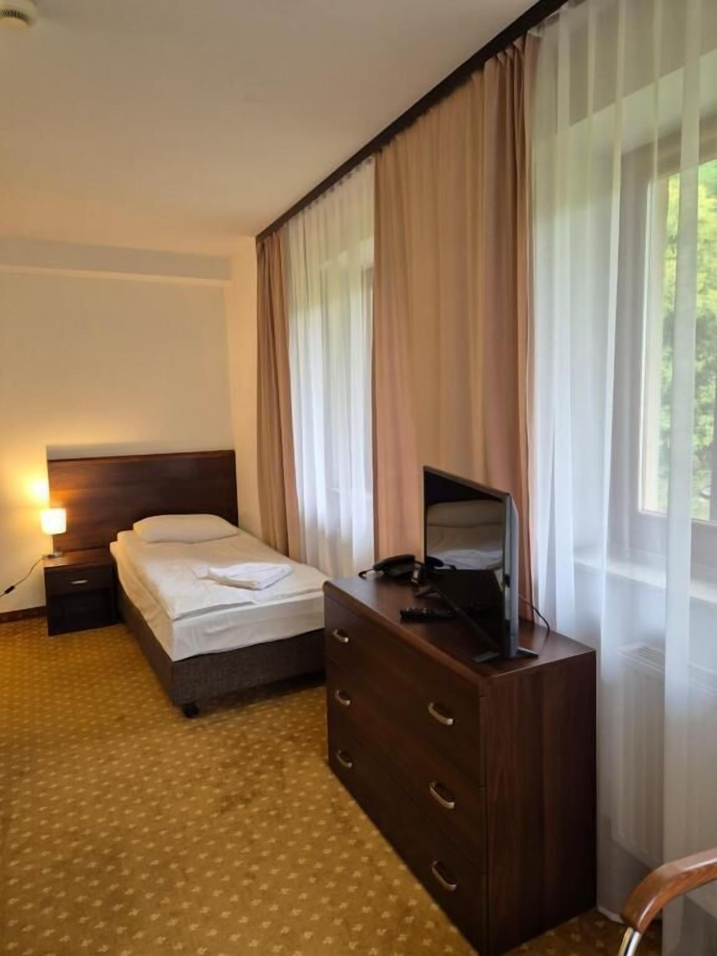 A hotel room with a bed, a dresser, a desk with a computer, a lamp, and a window with curtains.