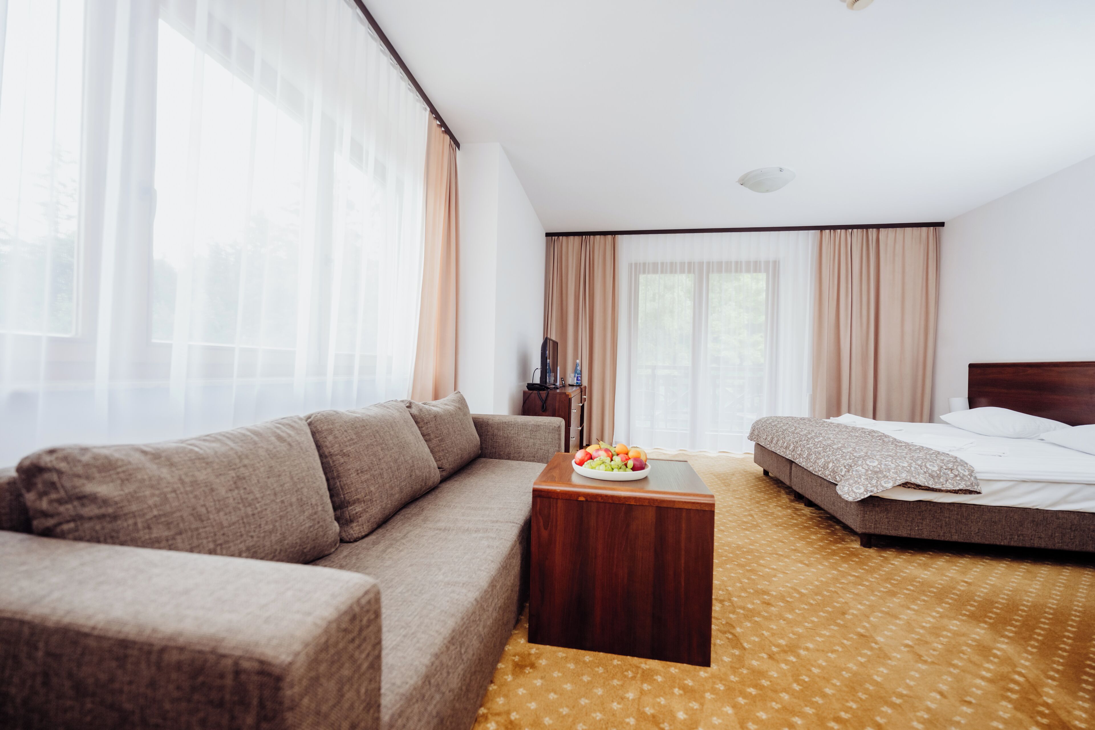 Quarto de hotel com sofá, cama, mesa de centro de madeira com frutas e janelas amplas com cortinas.