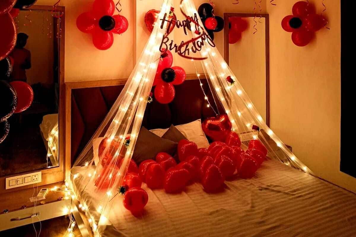 A room decorated with red and black balloons, a canopy with 'Happy Birthday' written on it, and a bed with red balloons and lights.