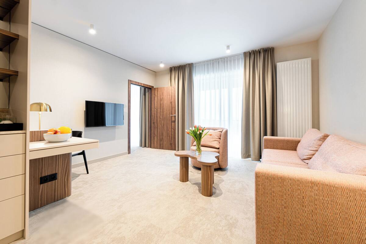 Ein modernes Hotelzimmer mit einer Couch, einem kleinen Tisch mit einer Vase, einem Schreibtisch mit einer Lampe und einem Fernseher an der Wand.