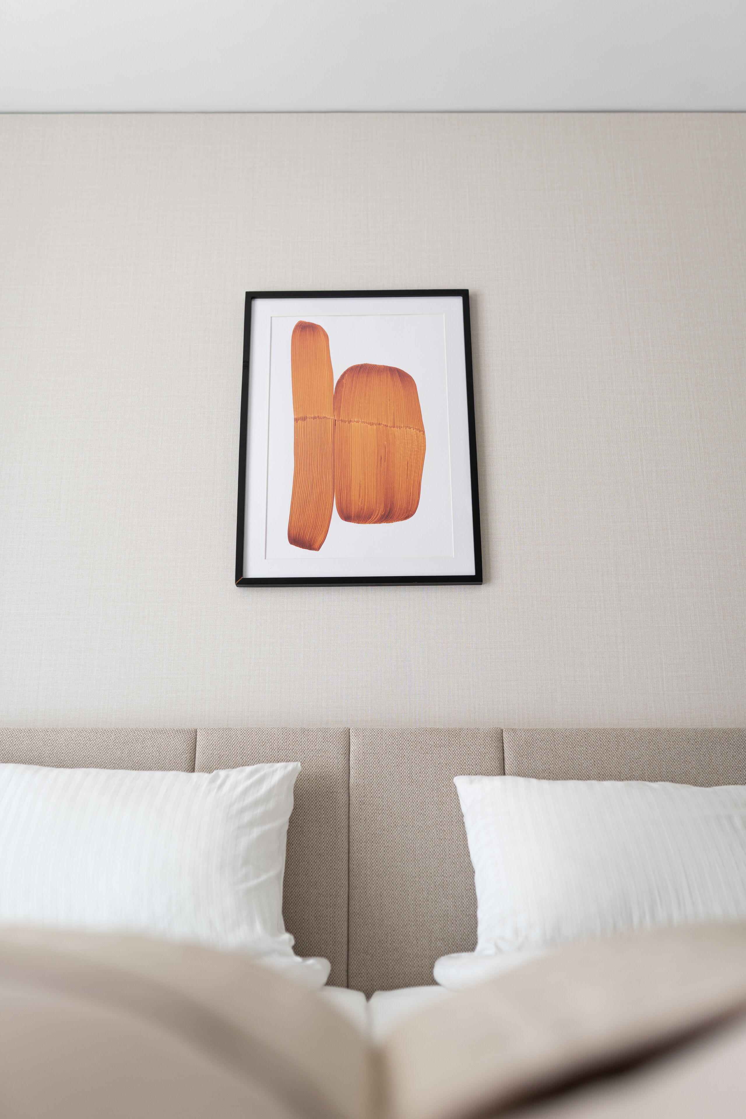 Una obra de arte enmarcada en una pared, sobre una cama con almohadas blancas y cabecero color crema.