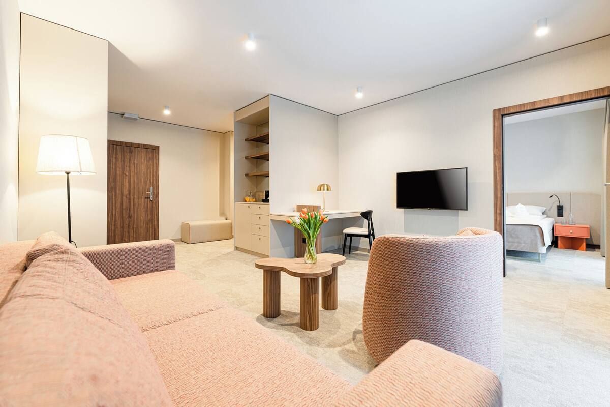 Ein modernes Hotelzimmer mit einer Couch, einem kleinen Holztisch mit einer Vase, einem Flachbildfernseher, einem Bett und einem Schreibtisch mit Stuhl.