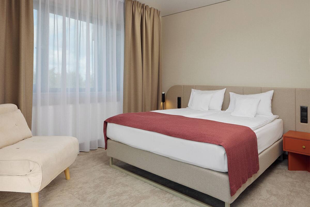 Ein Hotelzimmer mit einem Bett, einem Sessel, einem Nachttisch und einem an der Wand befestigten Lautsprecher.