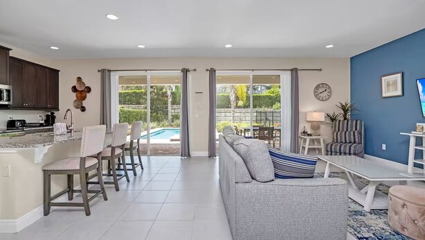 Una cocina moderna con armarios oscuros, un comedor con sofá gris y silla a rayas, y una piscina visible a través de puertas de vidrio.