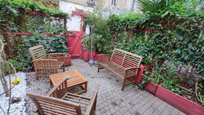 A patio with wooden furniture, including chairs and a table, surrounded by greenery and a red fence. - Jardin Secret Greenhouse (Ivry-sur-Seine)