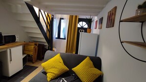 A compact living space with a kitchenette, a sofa with yellow cushions, a staircase, and a window with curtains. - Jardin Secret Greenhouse (Ivry-sur-Seine)