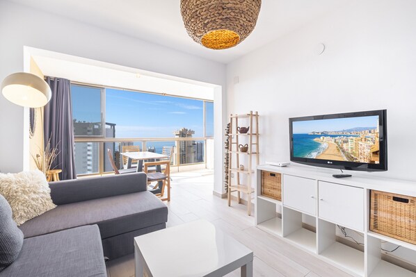 Una sala de estar moderna con un televisor grande, un sofá, una mesa de comedor y un balcón con vistas a la ciudad.