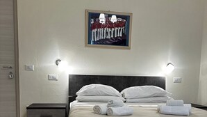 A neatly made bed with white linens and towels, a dark headboard, and a painting above it. There are two bedside tables with lamps, a door on the left, and a wooden floor. - Double Room with private bathroom in Terralba (Oristano) - Juniper (Terralba)