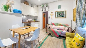 Een ruimtelijk ingerichte leefruimte met een eethoek, kitchenette en een bank met een 'JOLIE' kussen.