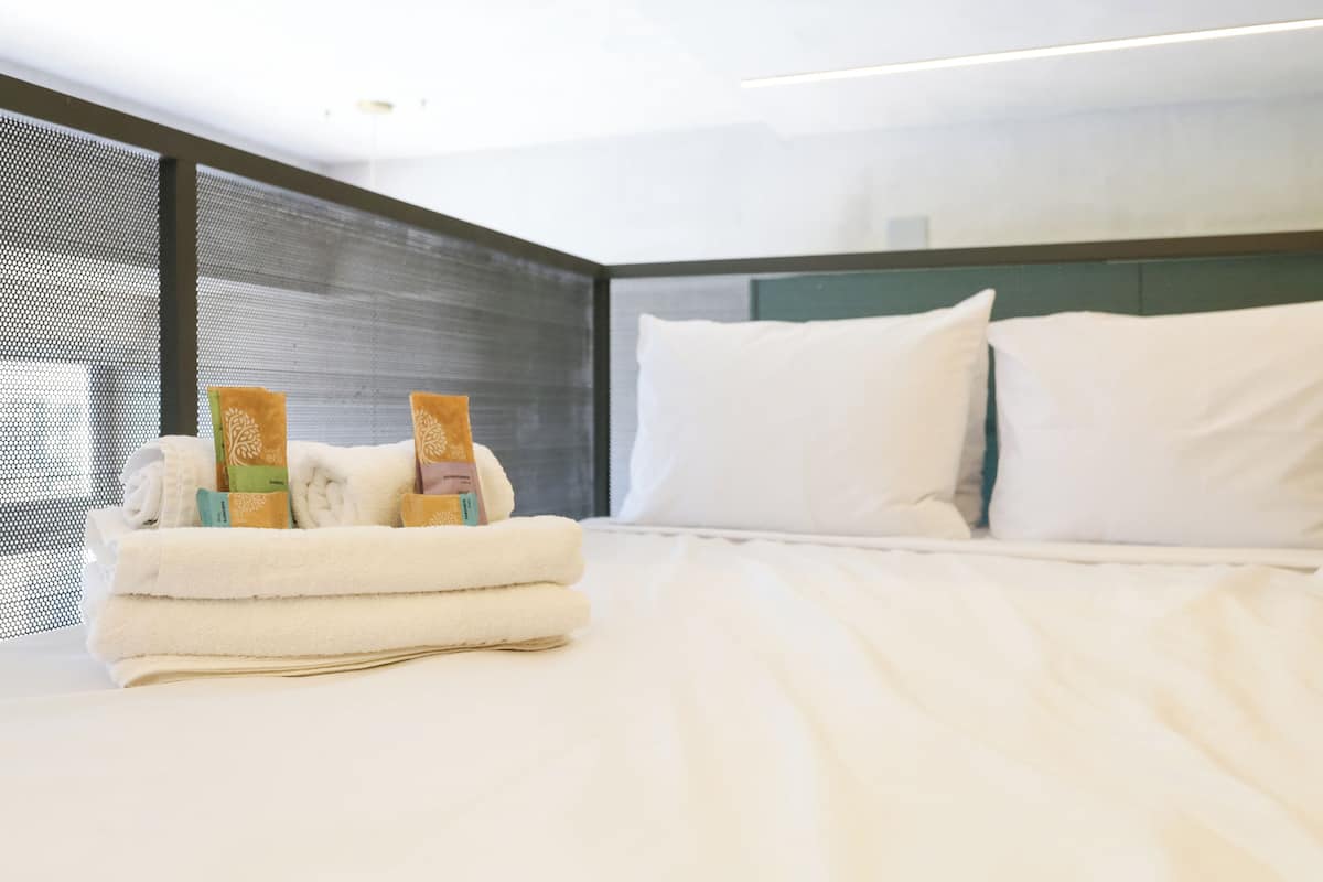 Una cama bien hecha con sábanas y almohadas blancas, una pila de toallas dobladas y una ventana con mosquitero.