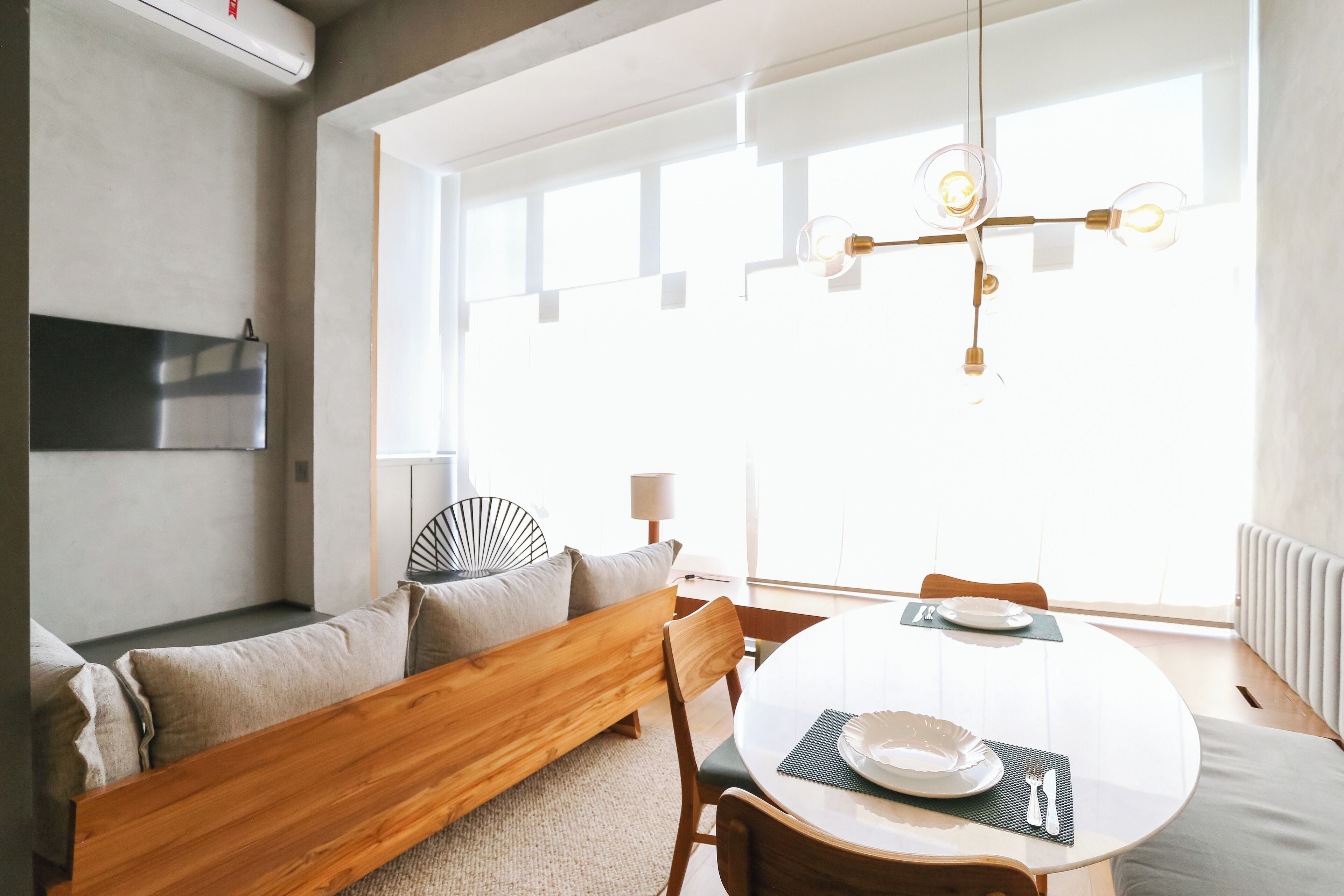 Un salon moderne avec un banc en bois, une table à manger ronde agrémentée d’assiettes et de couverts, et une grande fenêtre laissant entrer la lumière naturelle.