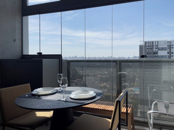 Une table à manger avec des assiettes et des verres, une chaise, et une vue sur les gratte-ciel d’une ville, visible à travers une fenêtre.