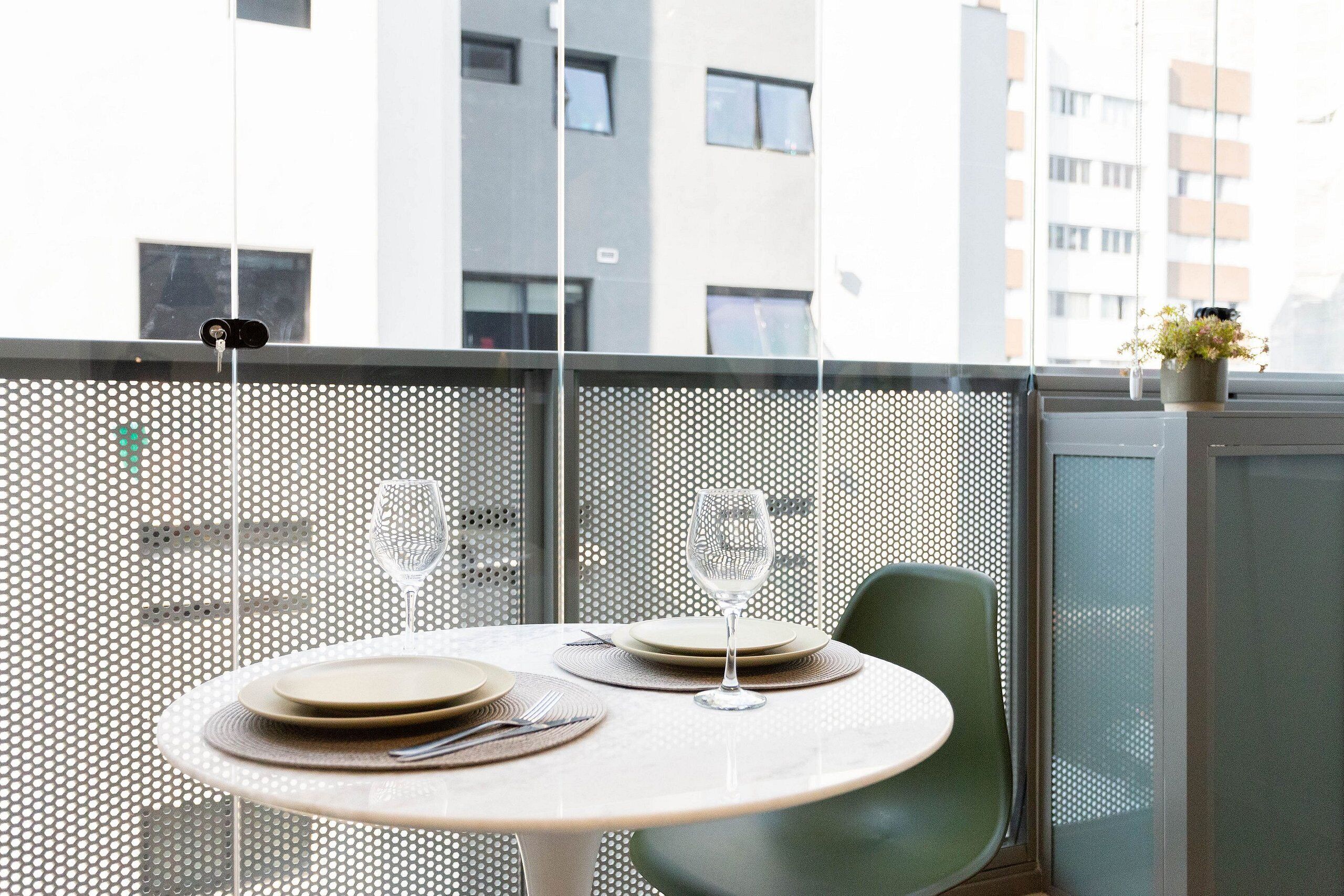 Un rincón de comedor con una mesa redonda dispuesta para dos, una silla verde y vistas a un edificio a través de un separador perforado.
