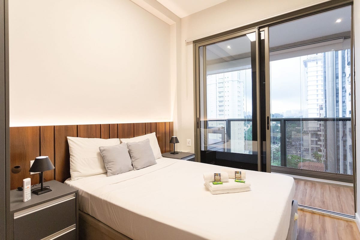 Una habitación de hotel moderna con una cama grande, una mesita de noche con lámpara, una pequeña mesa con un neceser y una puerta corrediza de vidrio que da a un balcón con vistas a la ciudad.