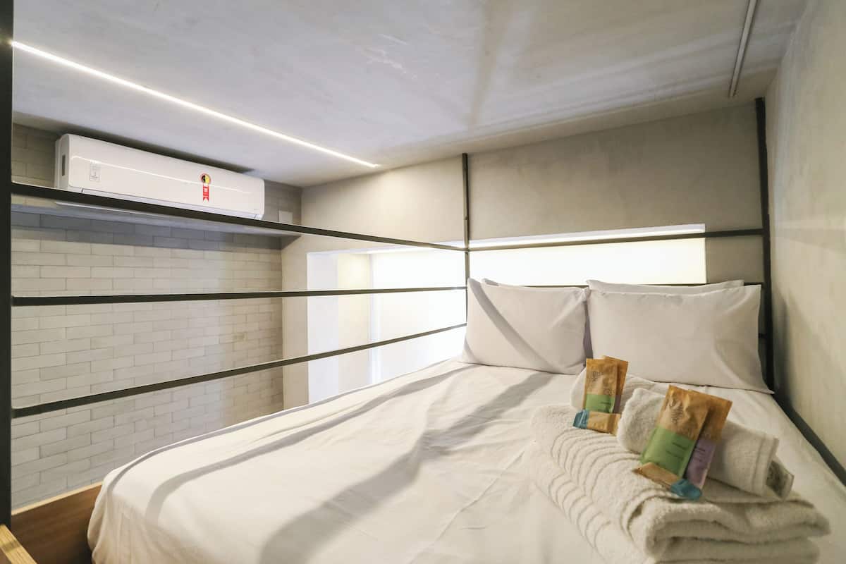 Una cama bien tendida con almohadas y una toalla doblada, un aire acondicionado de pared y un muro con diseño de ladrillo.