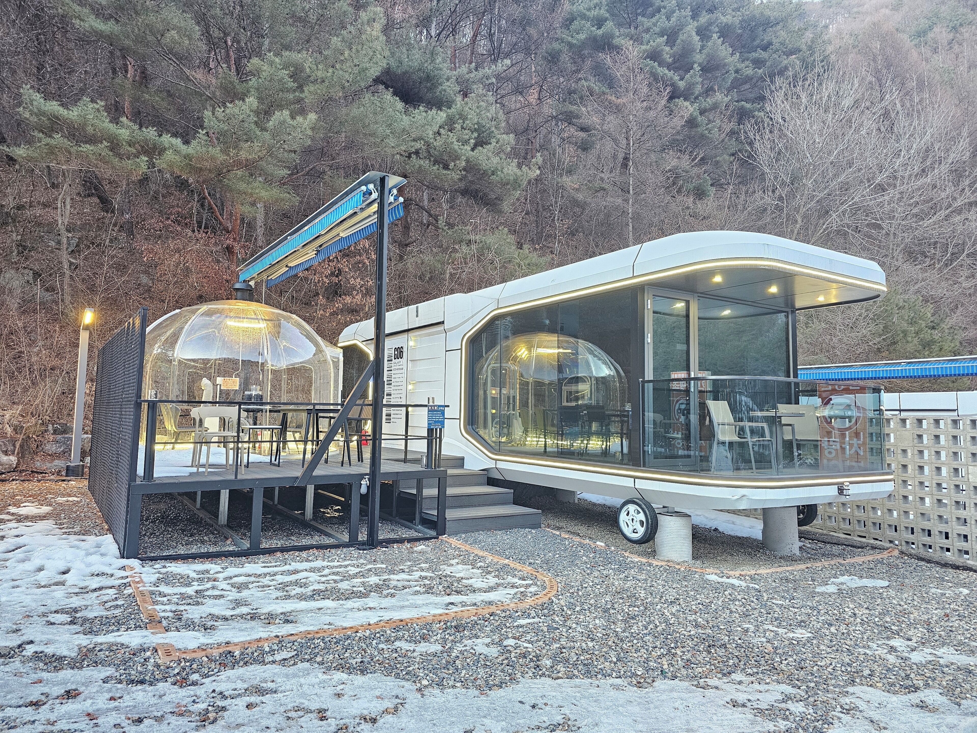 A modern RV with a transparent dome structure, a covered seating area, and a gravel ground.