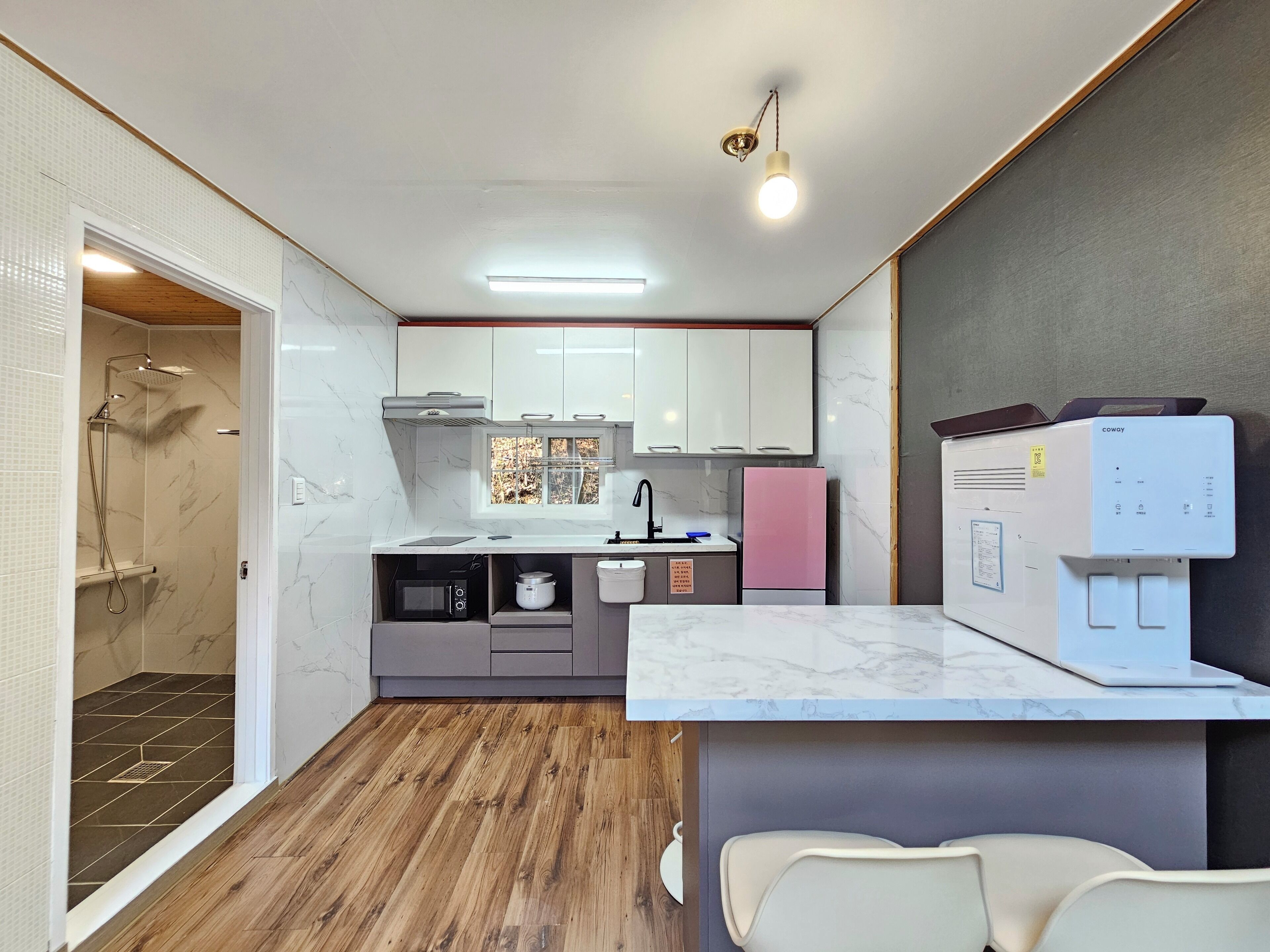 Una cocina compacta con microondas integrado, un refrigerador rosa y una ventana con vista a árboles.