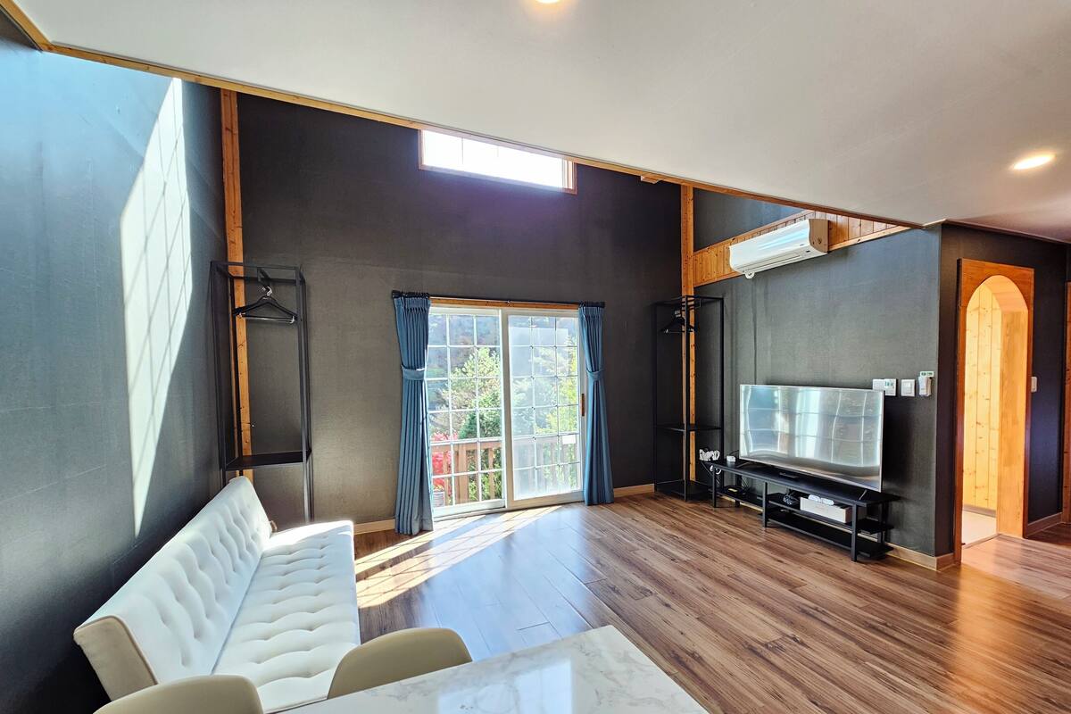 Una sala de estar moderna con un sofá blanco, una mesa de mármol, un televisor y un amplio ventanal con cortinas.