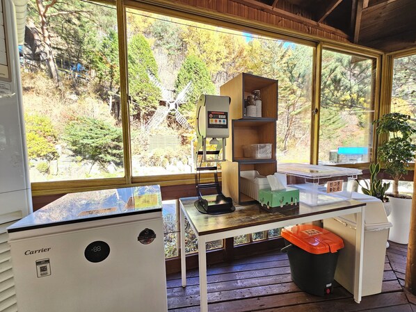 A room with a large window, a desk, a bookshelf, a trash bin, and a small refrigerator.