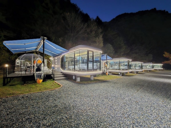 A row of transparent, futuristic bus-shaped structures with seating areas, set against a mountainous backdrop. - Galaxy x Hotel type Caravan Glamping&Pension (Pyeongchang)
