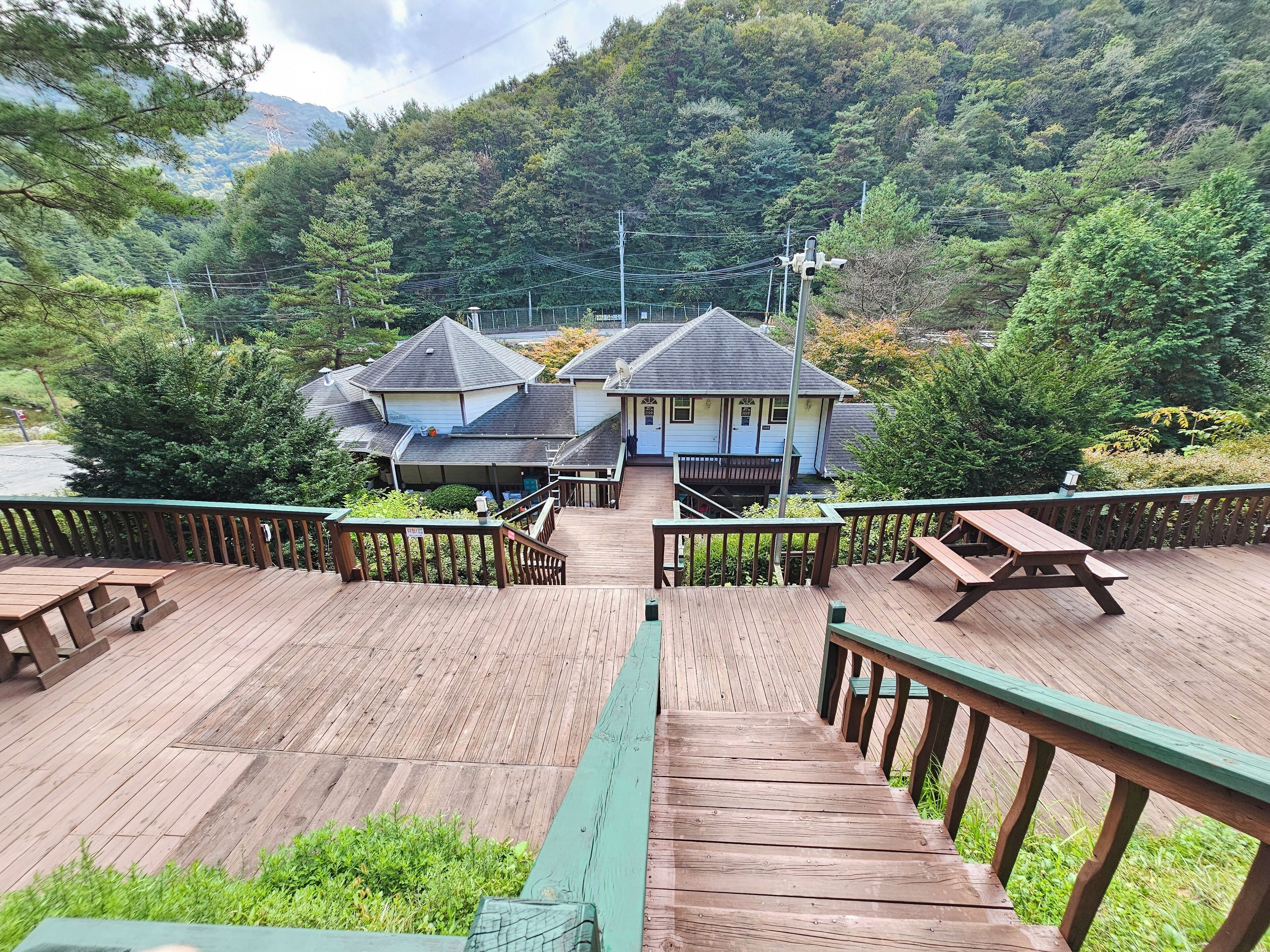 A wooden deck leading to a building with a covered patio area, surrounded by lush greenery and a mountainous backdrop.