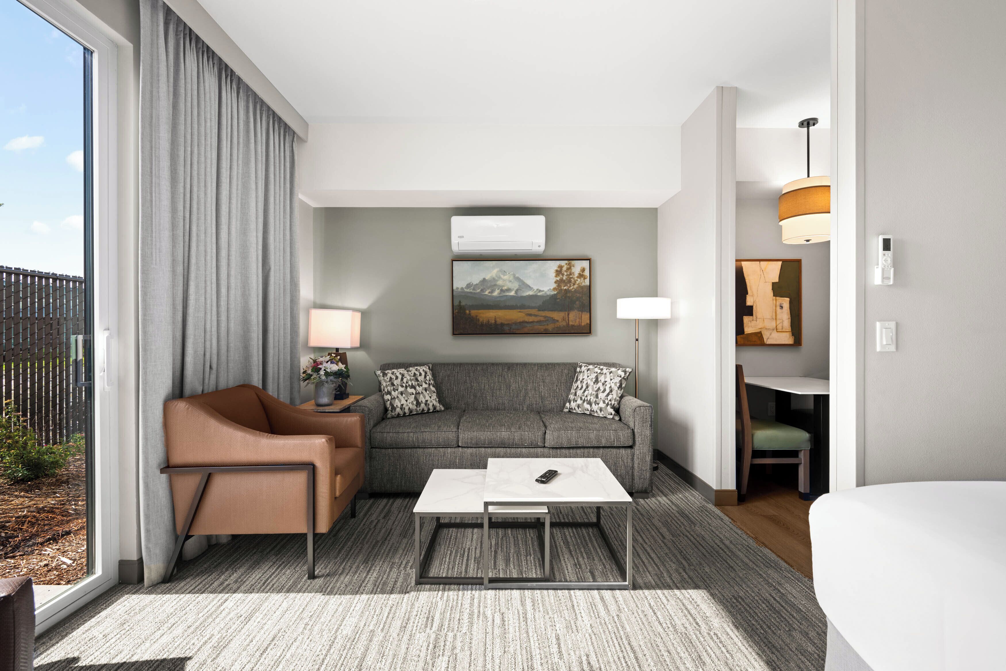 A modern hotel room with a grey sofa, a brown armchair, a white coffee table, and a large window with curtains.