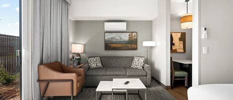 A modern hotel room with a grey sofa, a brown armchair, a white coffee table, and a large window with curtains.
