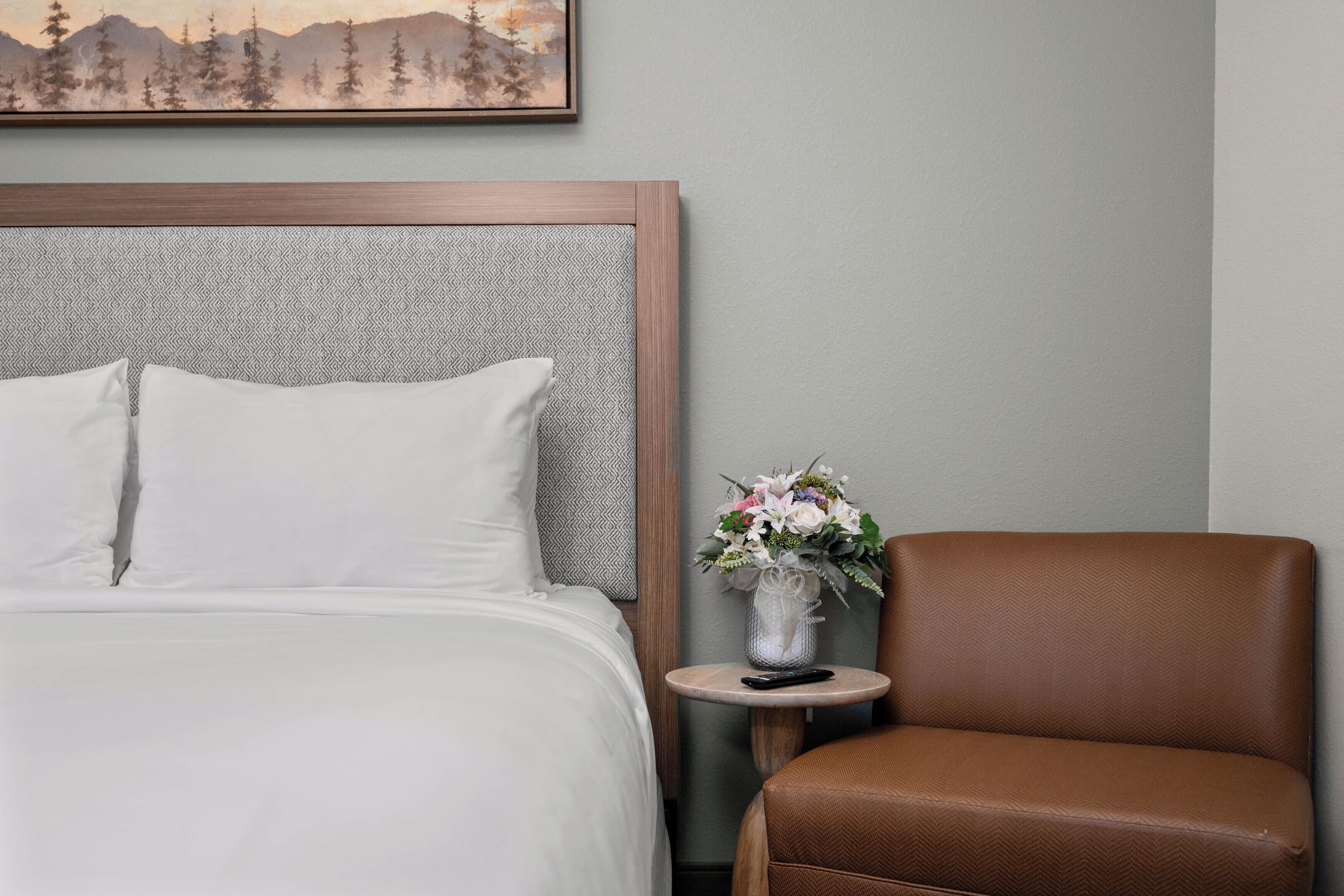 Een netjes opgemaakt bed met witte lakens, een klein rond tafeltje met een bloemenvaas, en een bruine leren fauteuil.