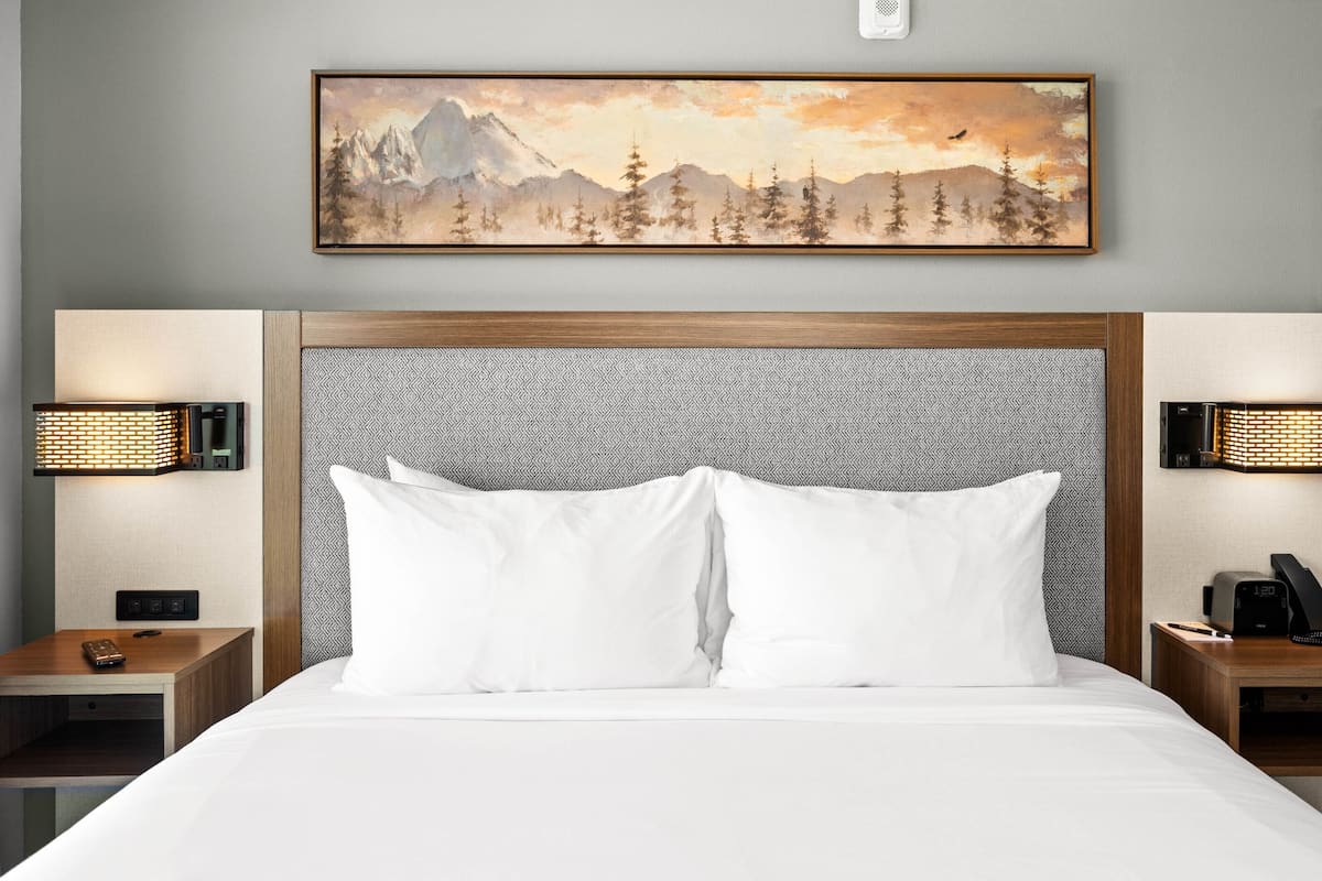 Ein Hotelzimmer mit einem großen Bett, zwei Nachttischen, einem Wandbild mit einer Berglandschaft und zwei Wandleuchten.