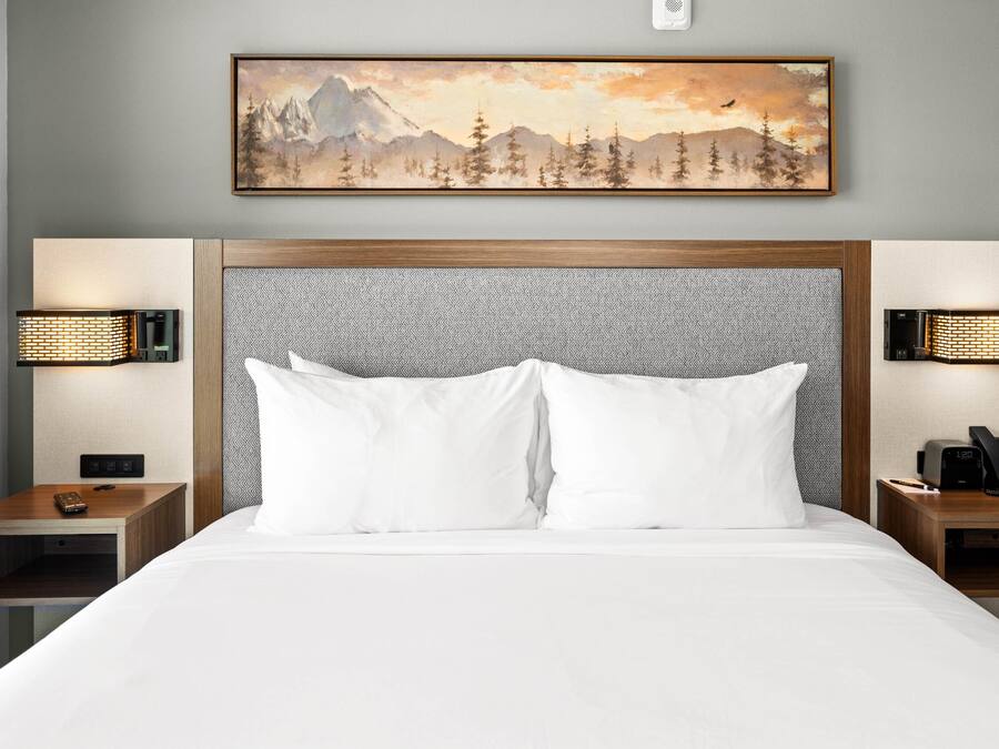 Quarto de hotel com uma cama grande, duas mesas de cabeceira, um quadro pendurado na parede com uma paisagem de montanhas e duas luminárias de parede.