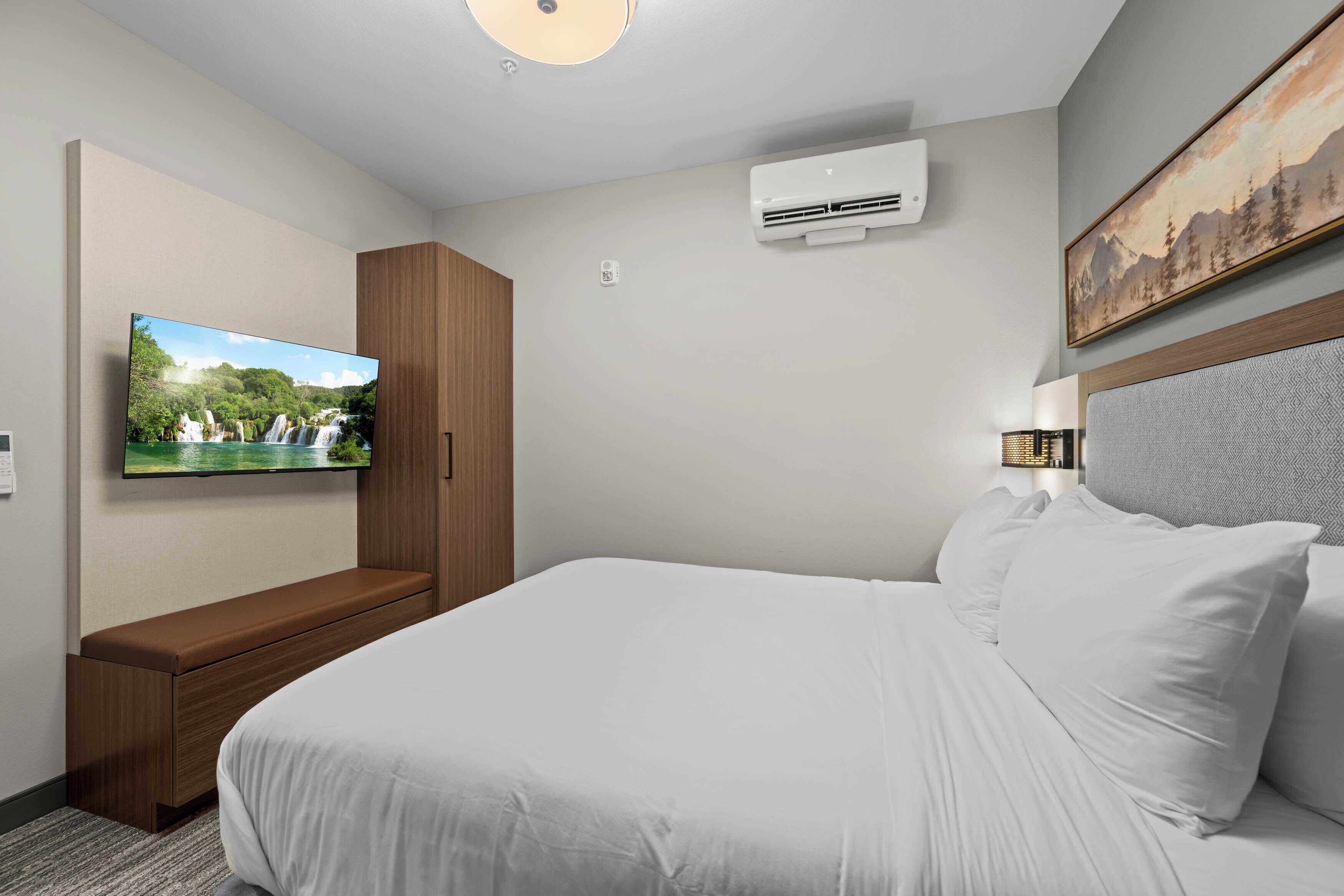 A hotel room with a bed, a TV mounted on the wall, a wardrobe, an air conditioning unit, and a painting above the bed.