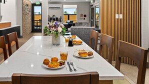 A hotel dining area with a long table, chairs, a vase with flowers, and breakfast items. - The Kulshan Hotel (Sedro-Woolley)