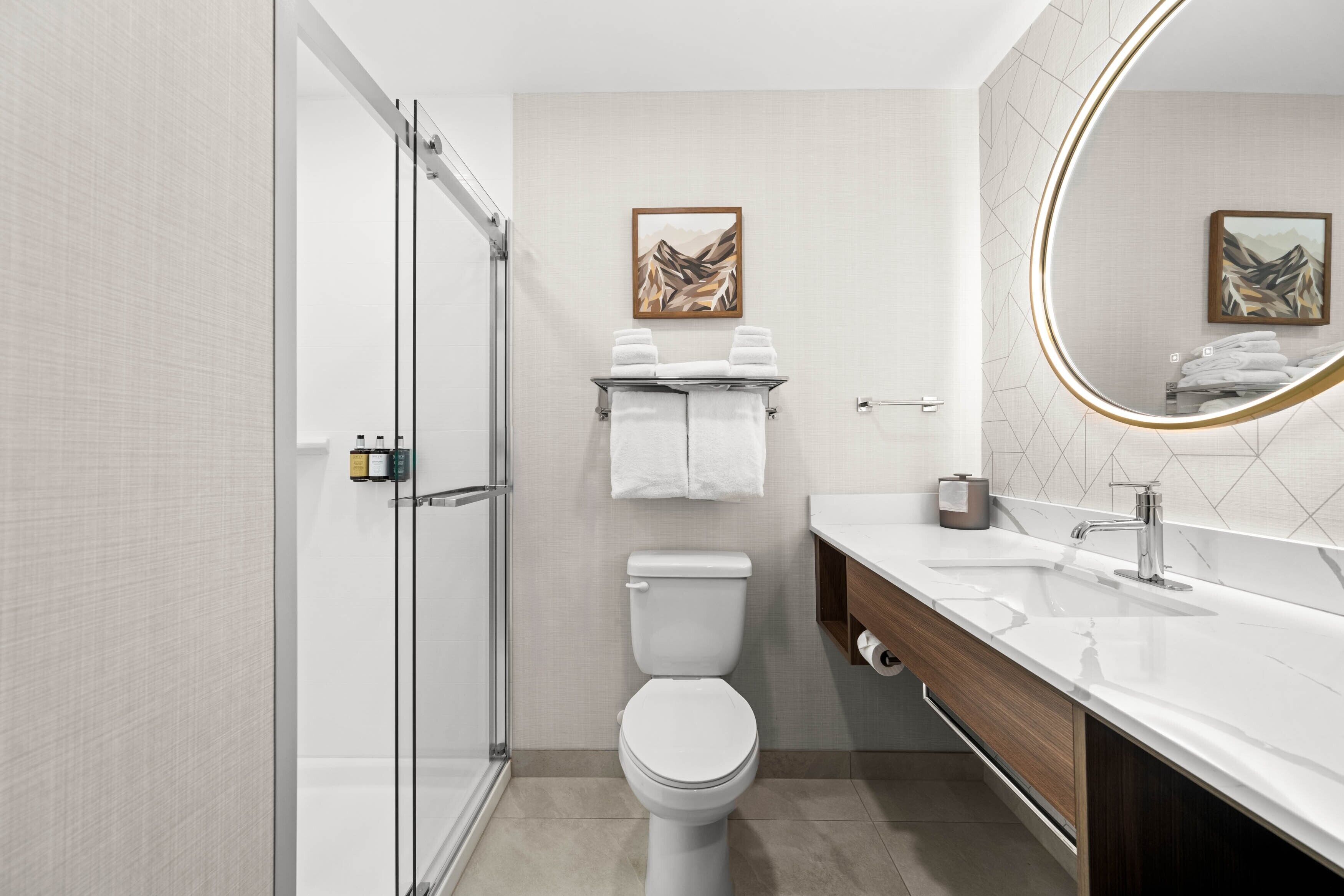 A modern bathroom with a glass shower enclosure, a white toilet, a sink with a mirror, and two towels on a shelf.