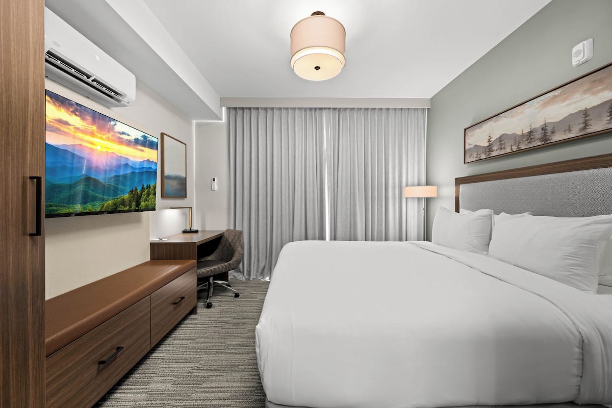 Ein Hotelzimmer mit einem großen Bett, einem Schreibtisch mit Stuhl, einem Flachbildfernseher, einer Deckenleuchte und einem Fenster mit Vorhängen.