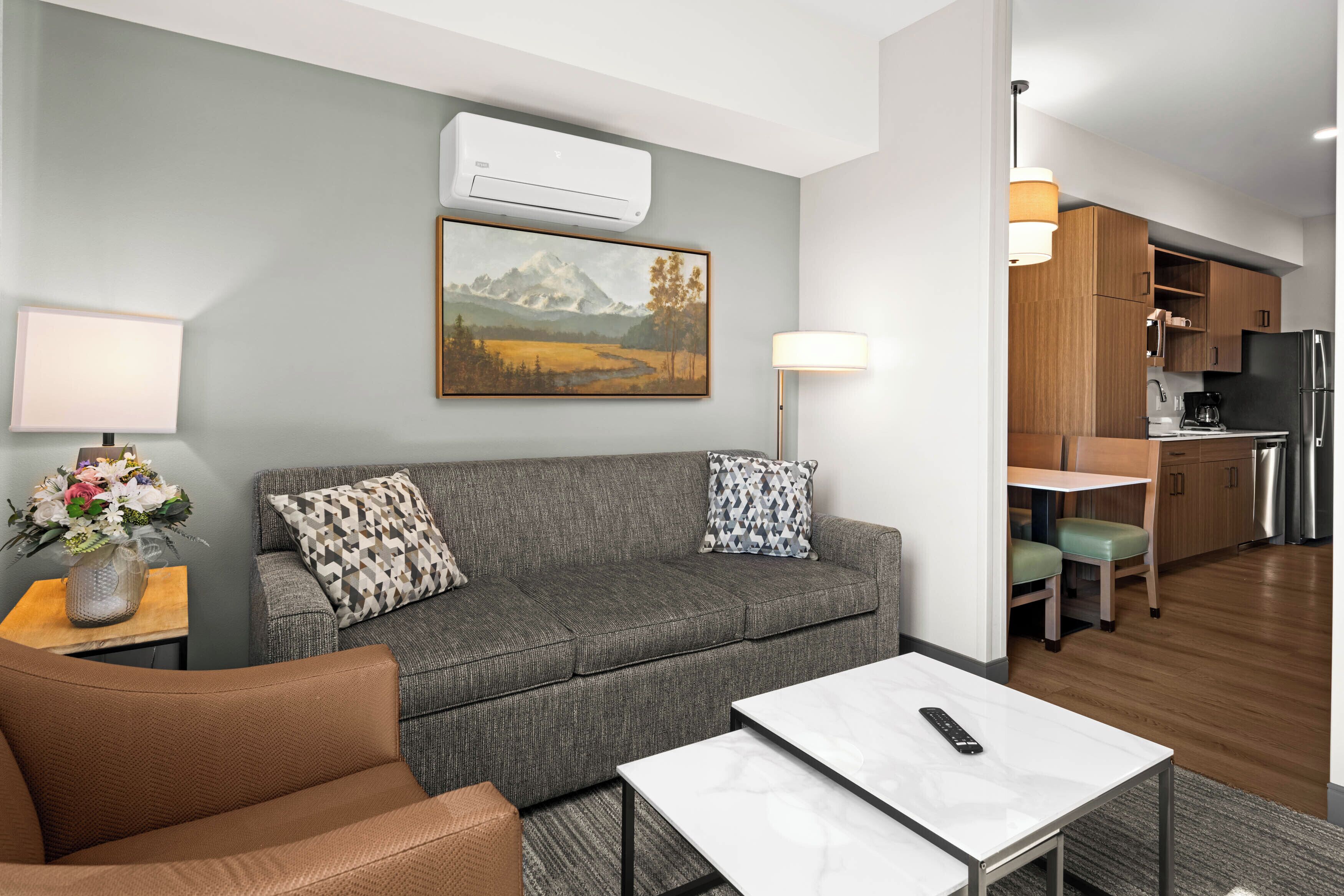 A modern living room with a grey sofa, a brown armchair, a white coffee table, and a painting of mountains on the wall.