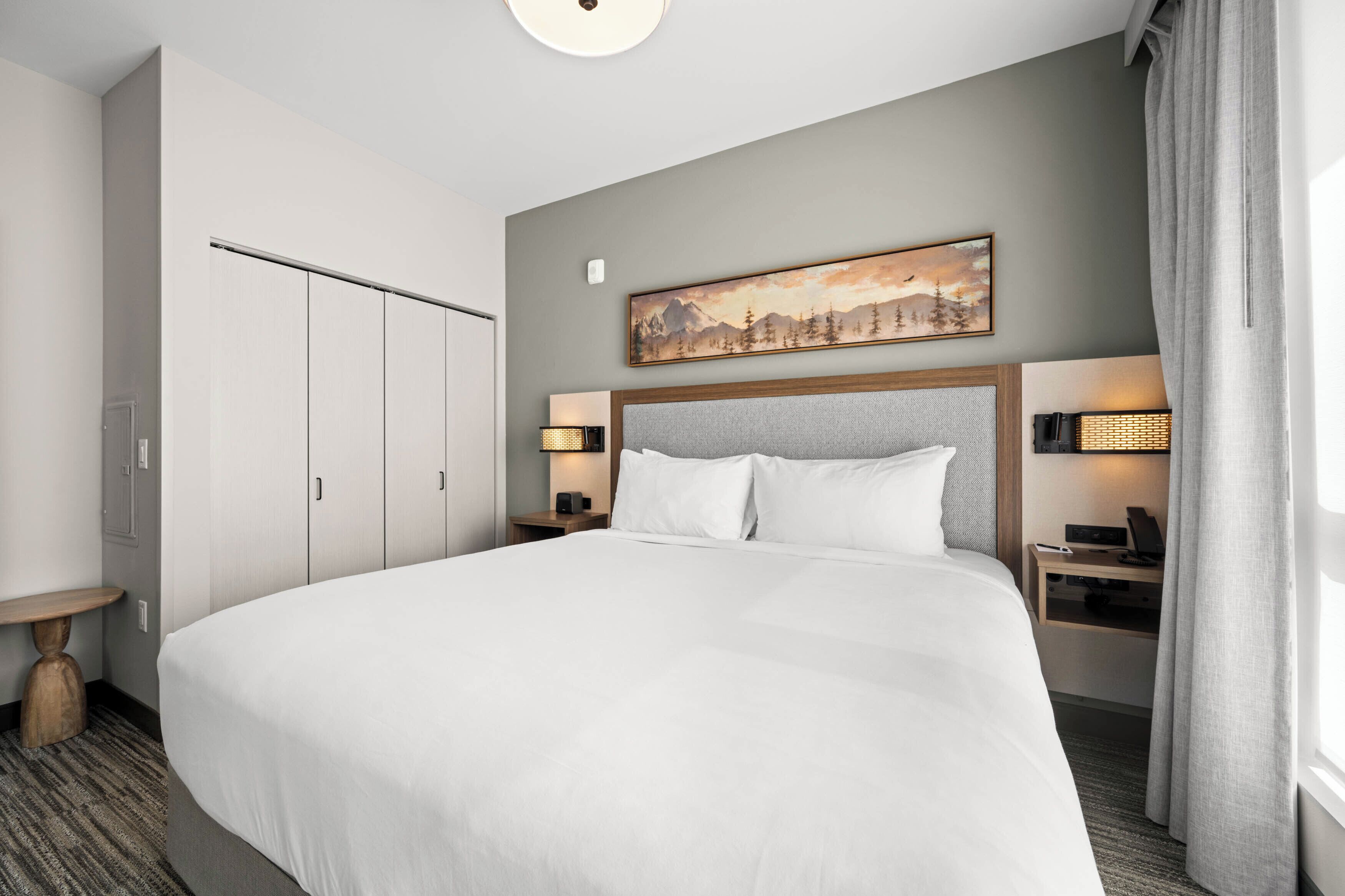 Habitación de hotel con una cama grande, un cuadro sobre la cama, dos lámparas de noche, un escritorio con lámpara y un armario.
