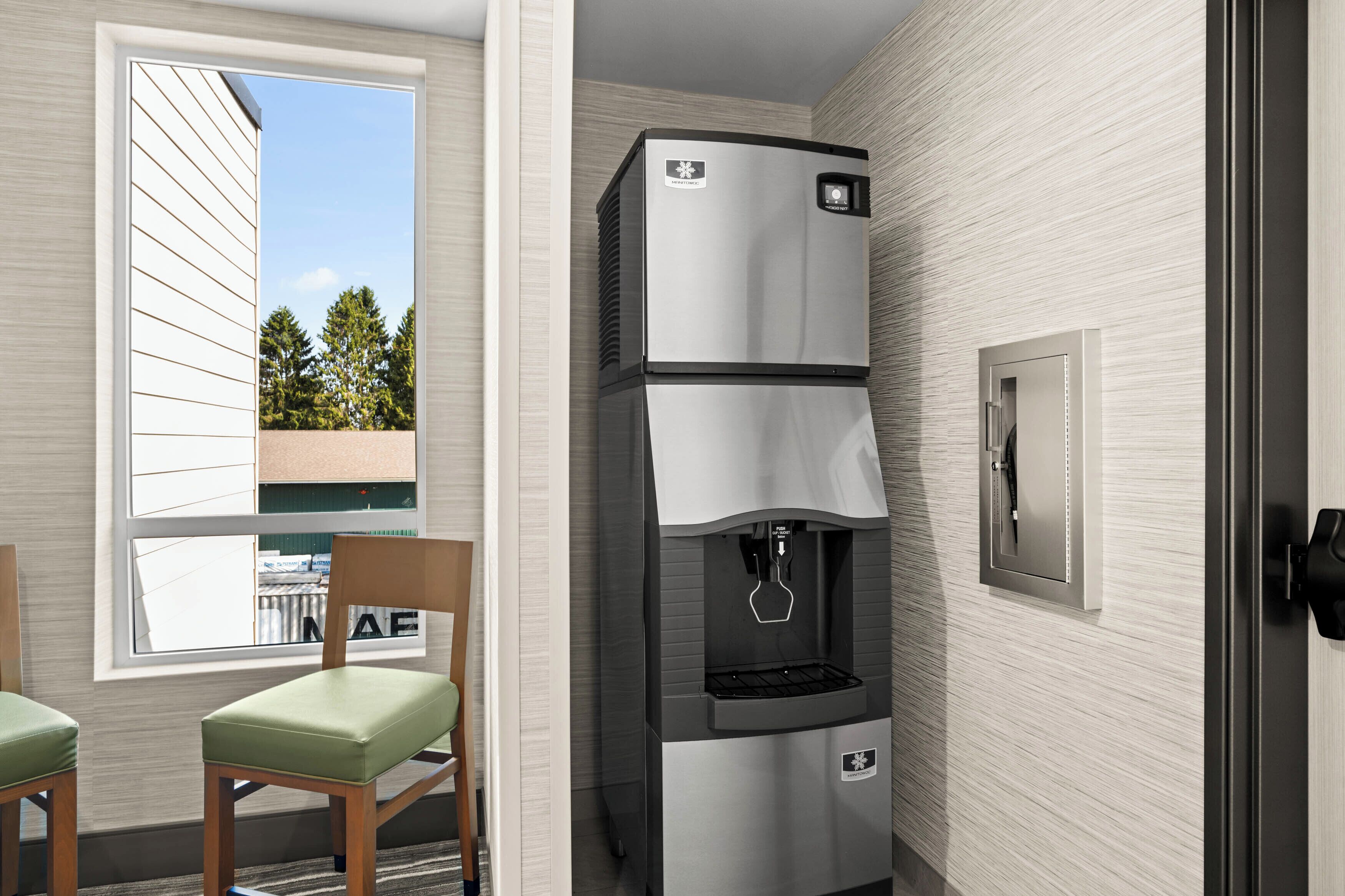 A compact kitchenette with a built-in refrigerator, a water dispenser, and a microwave. There is a window providing a view of trees and a building outside. Two chairs are placed in front of the window.