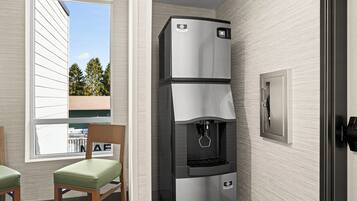 A compact kitchenette with a built-in refrigerator, a water dispenser, and a microwave. There is a window providing a view of trees and a building outside. Two chairs are placed in front of the window.